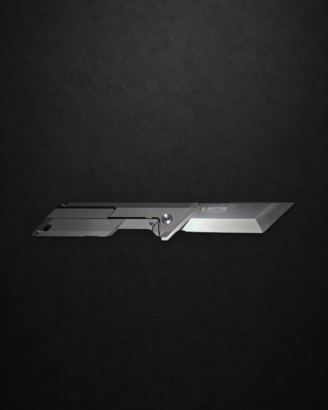A sleek, silver tactical folding knife with a minimalist design, displayed horizontally against a dark, textured black background. The brand name 'BAYONET' is visible on the blade.