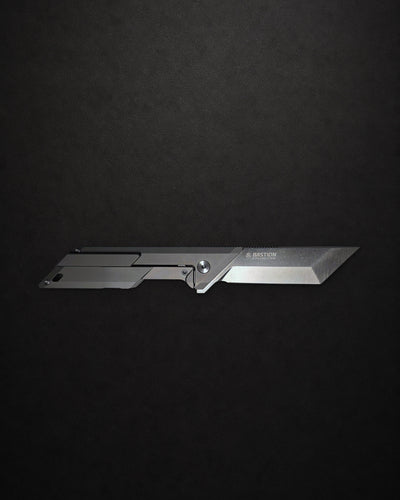 A sleek, silver tactical folding knife with a minimalist design, displayed horizontally against a dark, textured black background. The brand name 'BAYONET' is visible on the blade.